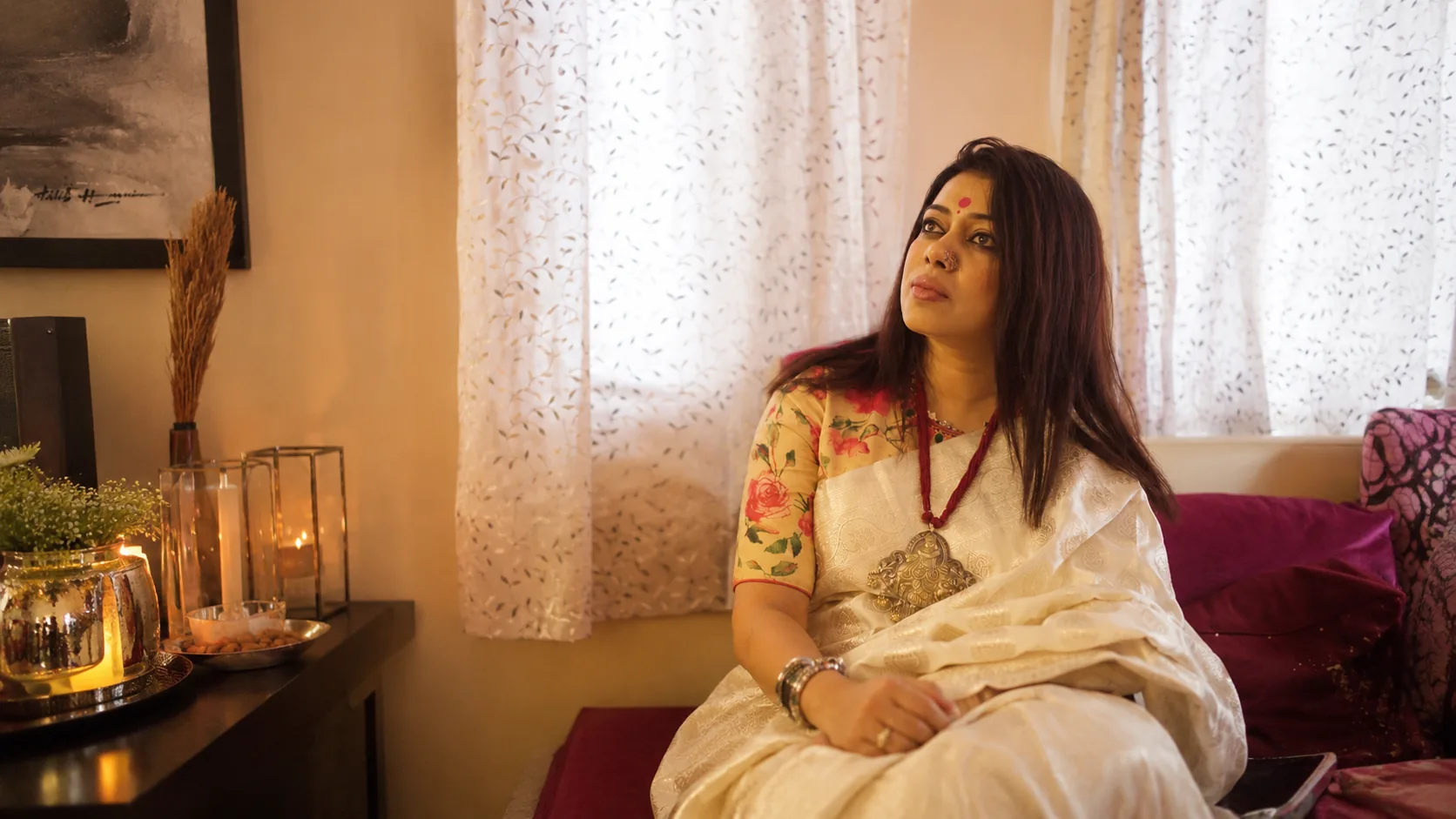 Barnali Chattopadhyay seated quietly at home in an ivory saree, candlelight beside her